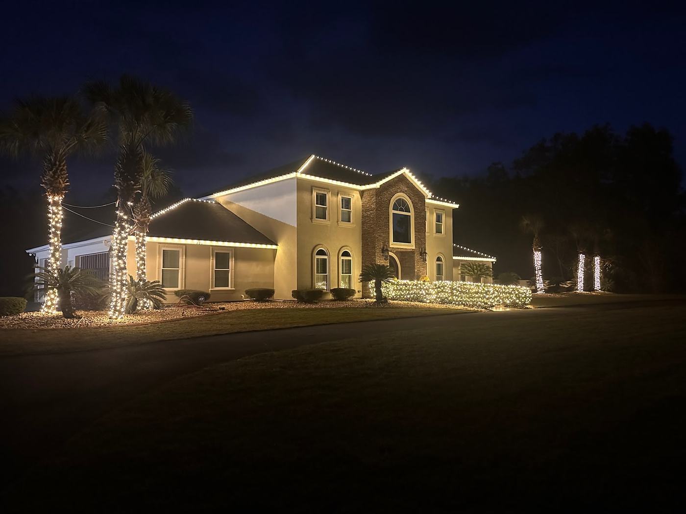 Warm white roofline lighting and wrapped palms on a Pensacola stucco home